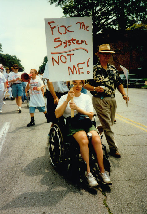 A protester who uses a wheelchair holds up a sign that says 'Fix The System: Not Me.'